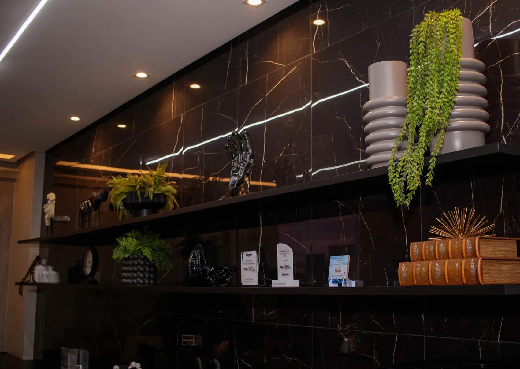 Parede de mármore preto da sede de Macapá, com iluminação linear, plantas e livros decorativos.
