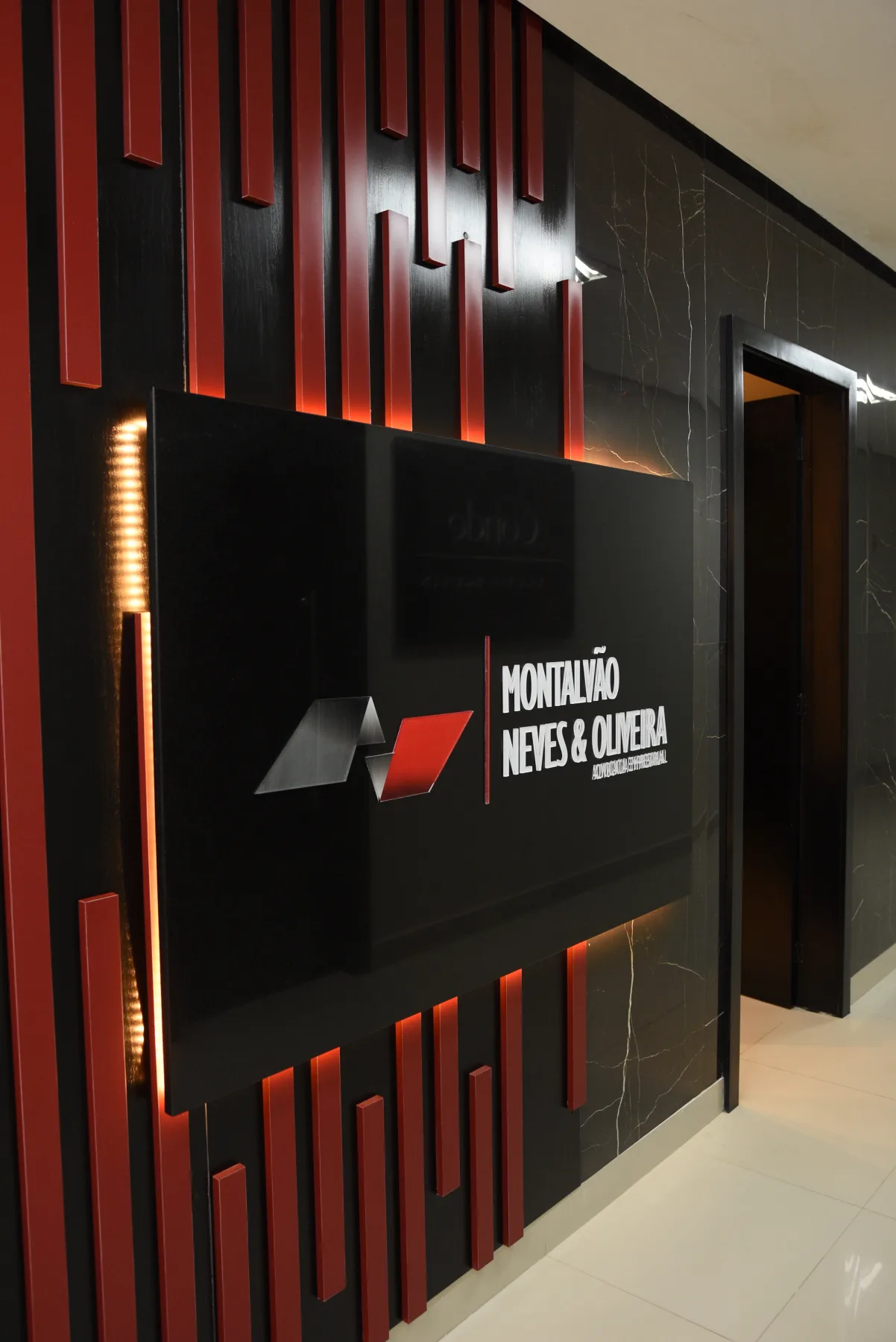 Parede da recepção da sede de Macapá em mármore preto e ripado vermelho, com o letreiro iluminado do Montalvão Neves & Oliveira.