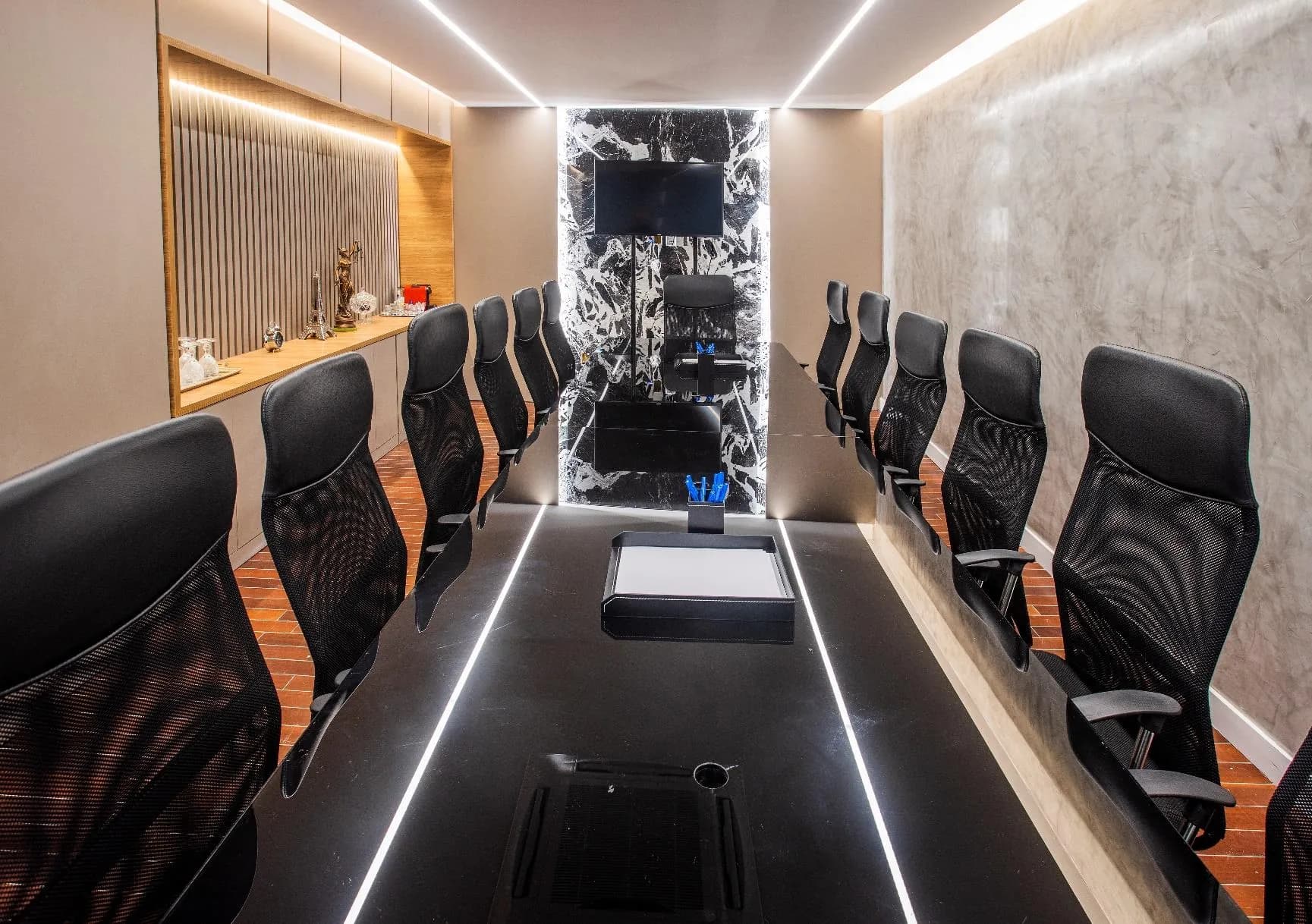 Sala de reunião com mesa preta alongada, painel de mármore ao fundo e televisor.